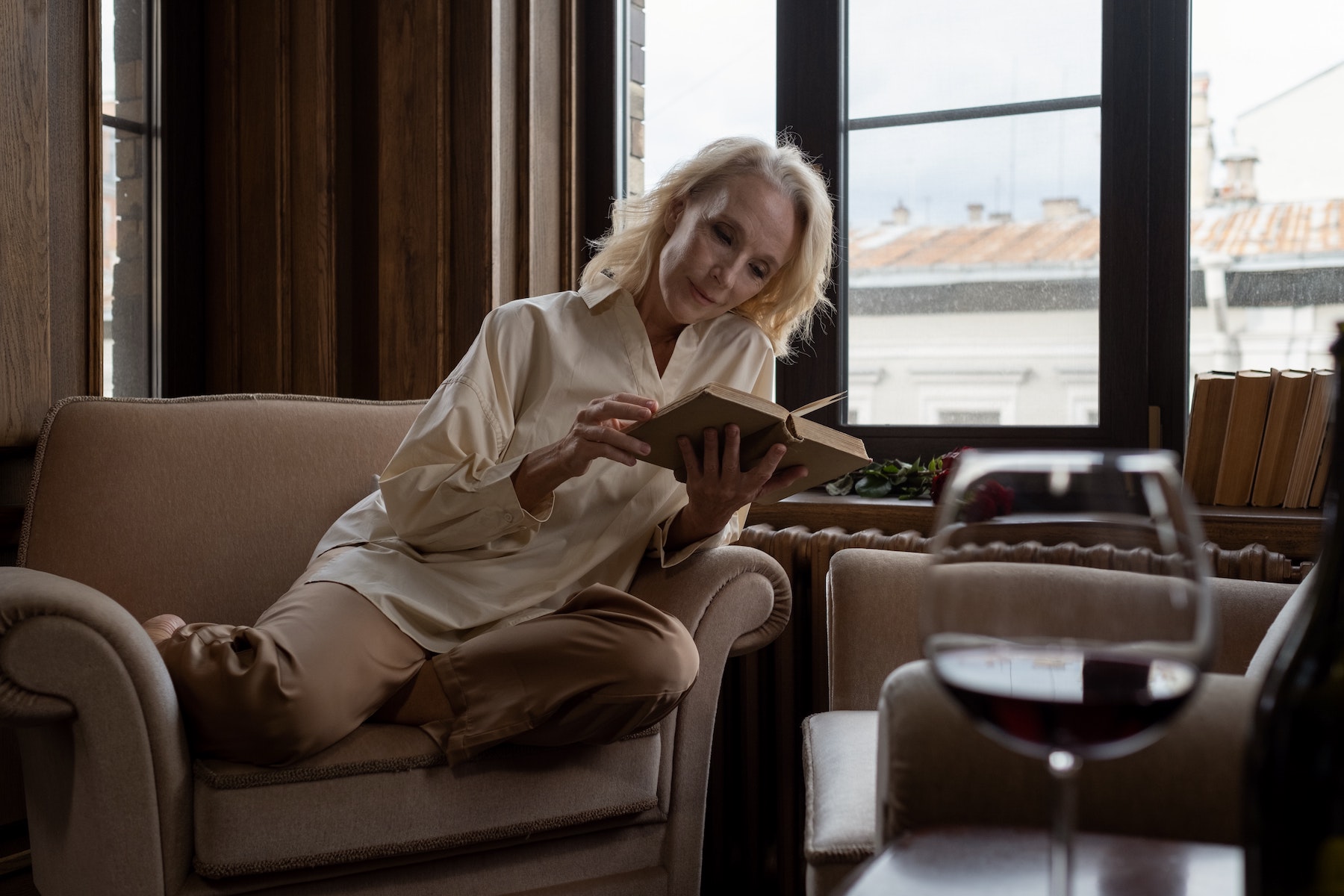 donna di mezza età legge un libro di poesie sul divano e ci abbina un calice di vino
